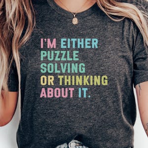 Puede incluir: Camiseta gris oscuro con el texto "I'M EITHER PUZZLE SOLVING OR THINKING ABOUT IT" en letras de colores. La camiseta es de cuello redondo y parece estar hecha de un material suave.