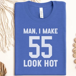 May include: A blue t-shirt with the text "MAN, I MAKE 55 LOOK HOT" in white block letters. The shirt is a crew neck style and is folded neatly. The shirt is made of a soft material.
