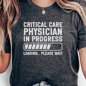 May include: Dark gray t-shirt with the text "CRITICAL CARE PHYSICIAN IN PROGRESS" in white, along with a loading bar graphic and the words "LOADING... PLEASE WAIT". The shirt is a crew neck.