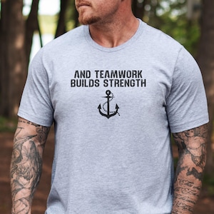 Puede incluir: Una camiseta gris con un ancla negra y el texto "AND TEAMWORK BUILDS STRENGTH".