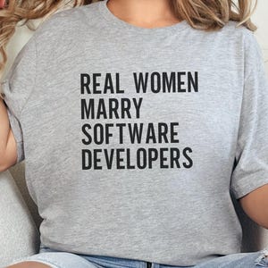 May include: A light gray t-shirt with the text "REAL WOMEN MARRY SOFTWARE DEVELOPERS" in black block letters. The shirt is a casual style, suitable for everyday wear.