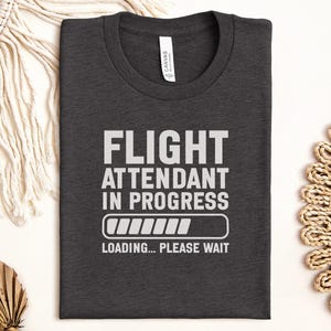 Puede incluir: Camiseta gris oscuro con el texto blanco "FLIGHT ATTENDANT IN PROGRESS". Debajo, un gráfico de barra de carga. La camiseta también incluye el texto "LOADING... PLEASE WAIT".