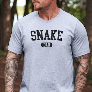 May include: A pale grey t-shirt featuring the text "SNAKE DAD" in black, bold capital letters. The word "DAD" is in a smaller, rectangular box below "SNAKE". The shirt has short sleeves.