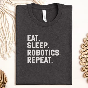 May include: Charcoal grey t-shirt featuring the text "EAT. SLEEP. ROBOTICS. REPEAT." in white. The shirt has a crew neck and a "CANVAS" label. It is folded and displayed on a white surface, accompanied by decorative elements.