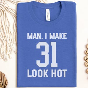 May include: Royal blue t-shirt with the text "MAN, I MAKE 31 LOOK HOT" in white block letters. The shirt is a crew neck style and is folded neatly. The shirt is made of a soft material.