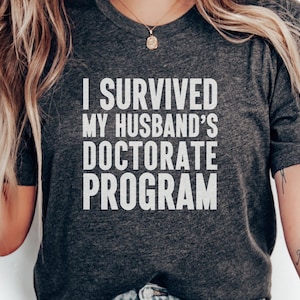 Könnte beinhalten: Ein graues T-Shirt mit weißem Text, der "Ich habe das Doktorat meines Mannes überlebt" lautet.