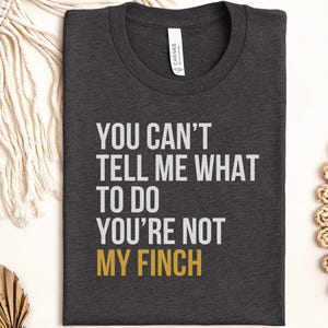May include: A dark charcoal grey crew neck t-shirt featuring the white text "YOU CAN'T TELL ME WHAT TO DO YOU'RE NOT MY FINCH" with the word "FINCH" printed in a mustard yellow colour. The shirt has a standard short-sleeve design and a neck label.