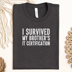 May include: Dark gray t-shirt with the text "I SURVIVED MY BROTHER'S IT CERTIFICATION" in white block letters. The shirt has a crew neck and a tag that says "CANVAS". The shirt is folded and lying on a white surface.
