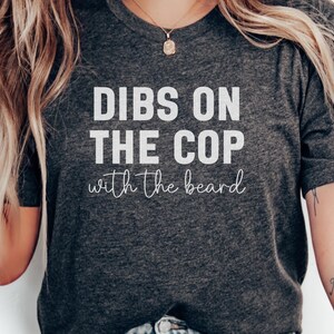 May include: A charcoal grey t-shirt featuring the white text "DIBS ON THE COP with the beard". The shirt has a crew neck and short sleeves. The text is in a bold, sans-serif font.