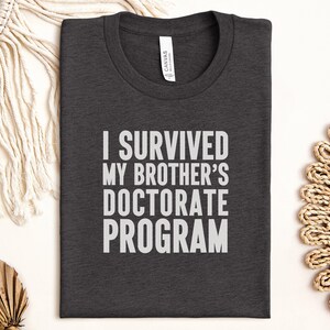 Puede incluir: Camiseta gris oscuro con el texto "I SURVIVED MY BROTHER'S DOCTORATE PROGRAM" en letras blancas. La camiseta está hecha de un material suave y tiene cuello redondo. La camiseta está doblada cuidadosamente sobre una superficie blanca.