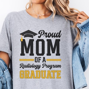 May include: A gray t-shirt with the text "Proud MOM of a Radiology Program GRADUATE" in black and gold lettering. The design includes a graduation cap. The shirt is a casual style.