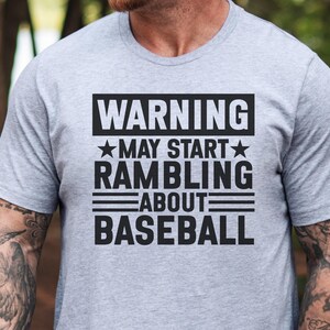 May include: A pale grey t-shirt featuring the black text "WARNING MAY START RAMBLING ABOUT BASEBALL". The text is in a rectangular block. The shirt has short sleeves and a crew neck.