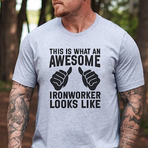 May include: A light grey t-shirt with the text "THIS IS WHAT AN AWESOME IRONWORKER LOOKS LIKE" in black. The design features two thumbs-up gestures. The shirt is made of a soft material.