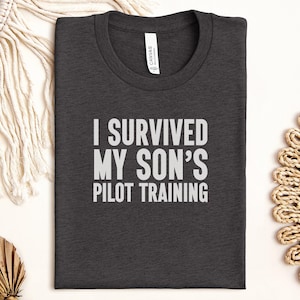 May include: Dark grey t-shirt with the white text "I SURVIVED MY SON'S PILOT TRAINING". The t-shirt is folded and has a crew neck. The fabric appears soft. The shirt is a crew neck style.