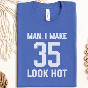 May include: A blue t-shirt with the text "MAN, I MAKE 35 LOOK HOT" in white block letters. The shirt is folded and lies on a neutral surface, with decorative elements in the background.