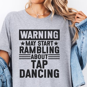 Puede incluir: Camiseta gris claro con el texto negro "WARNING MAY START RAMBLING ABOUT TAP DANCING". El texto está dispuesto en un bloque rectangular con acentos de estrella. La camiseta tiene mangas cortas y cuello redondo.