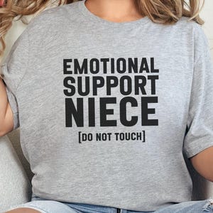 May include: Gray t-shirt with the text "Emotional Support Niece [Do Not Touch]" printed in black.