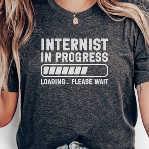 May include: Dark gray t-shirt with the text "INTERNIST IN PROGRESS" in white, along with a loading bar graphic. Below the graphic, the text reads "LOADING... PLEASE WAIT". The shirt is a crew neck.