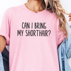 May include: A light pink t-shirt with the text "CAN I BRING MY SHORTHAIR?" in black, casual font. The shirt has short sleeves and is worn with a denim jacket.