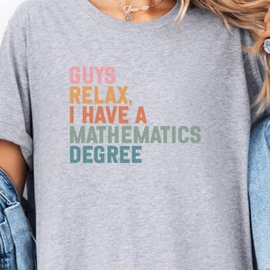May include: A light grey t-shirt featuring the text "GUYS RELAX, I HAVE A MATHEMATICS DEGREE" in a retro colour block design. The text is in pink, orange, green, and teal. The shirt has rolled-up sleeves.