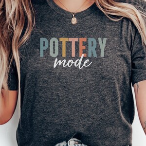 May include: A dark grey t-shirt with the text "POTTERY mode" in colorful letters. The letters are in shades of blue, yellow, pink, and green.