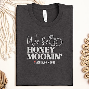 May include: Dark gray t-shirt with the text "We be Honey Moonin'" in white script and block letters, along with a diamond ring graphic. Below the text is "Aspen, CO • 2026" in white. The shirt is folded and lies on a white surface.