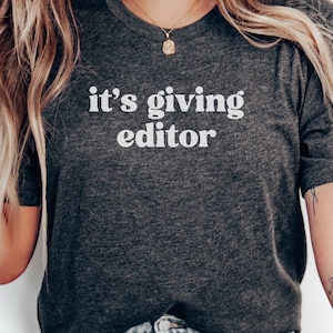 May include: Dark gray t-shirt with the text "it's giving editor" in white block letters. The shirt is a crew neck style and appears to be made of a soft material. A gold necklace is visible at the neckline.