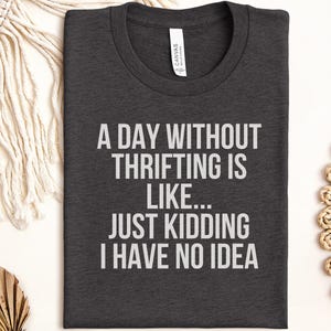Könnte beinhalten: Ein dunkel anthrazitfarbenes T-Shirt mit Rundhalsausschnitt und weißem Text auf der Vorderseite, der lautet: A DAY WITHOUT THRIFTING IS LIKE... JUST KIDDING I HAVE NO IDEA. Das T-Shirt hat einen Standardkragen und kurze Ärmel aus Stoff.