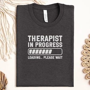 May include: Dark gray t-shirt with the text "THERAPIST IN PROGRESS" in white, along with a loading bar graphic and the words "LOADING... PLEASE WAIT". The shirt is made of a soft material.