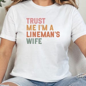Op de afbeelding: Wit T-shirt met de tekst "TRUST ME I'M A LINEMAN'S WIFE" in een retro kleurenschema. De tekst is verticaal gestapeld. Het shirt heeft korte mouwen en lijkt van een zacht materiaal te zijn.