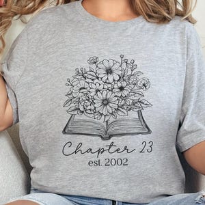 Puede incluir: Camiseta gris claro con un diseño floral en blanco y negro sobre una ilustración de un libro abierto. El texto "Chapter 23 est. 2002" está impreso debajo. La camiseta es de estilo casual.