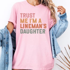 Op de afbeelding: Zachtroze T-shirt met de tekst "TRUST ME I'M A LINEMAN'S DAUGHTER" in een retro kleurenschema. De tekst is in roze, oranje en groen op elkaar gestapeld. Het T-shirt heeft een ronde hals.