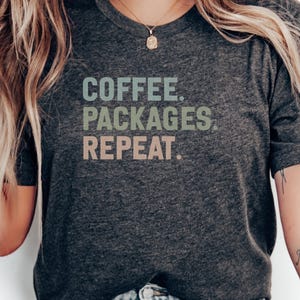 May include: A charcoal grey t-shirt featuring the words "COFFEE. PACKAGES. REPEAT." in a layered, muted colour scheme. The shirt has short sleeves and a crew neck. The text is in a sans-serif font.
