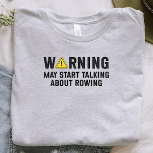 May include: Pale grey t-shirt featuring the text "WARNING MAY START TALKING ABOUT ROWING" in black, with a yellow hazard symbol. The shirt is displayed on a white surface, with a green plant and denim jeans visible.