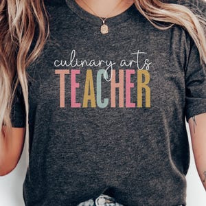 May include: Dark gray t-shirt with the words "culinary arts TEACHER" in colorful block letters. The text is in white, pink, blue, and yellow. The shirt is made of a soft material.