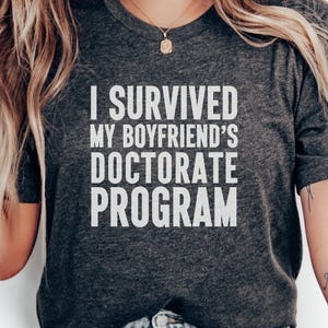 May include: Dark grey t-shirt with the white text "I SURVIVED MY BOYFRIEND'S DOCTORATE PROGRAM." The shirt is a crew neck and has short sleeves. A gold necklace is visible.