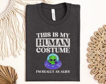 May include: Dark gray t-shirt with the text "THIS IS MY HUMAN COSTUME" above a cartoon alien in a spaceship, and the text "I'M REALLY AN ALIEN" below. The alien is green, the spaceship is purple and blue, and the text is white.