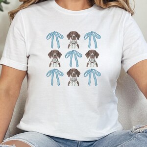 May include: White t-shirt featuring a repeating pattern of brown and white dogs with blue bows. The dogs have brown spots and are wearing collars. The bows are light blue. The shirt is a casual style.