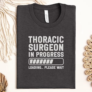 May include: Dark gray t-shirt with the text "THORACIC SURGEON IN PROGRESS" in white, along with a loading bar graphic and the words "LOADING... PLEASE WAIT." The shirt is made of a soft material and has a crew neck.