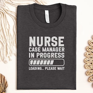 May include: Dark gray t-shirt with the text "NURSE CASE MANAGER IN PROGRESS" in white block letters. Below the text is a loading bar graphic and the words "LOADING... PLEASE WAIT". The shirt is made of a soft material.