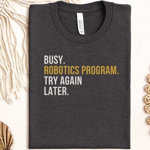 May include: A dark charcoal gray crew neck t-shirt featuring the white and gold text BUSY. ROBOTICS PROGRAM. TRY AGAIN LATER. The shirt has a standard short-sleeve design with a visible brand label inside the collar area on the inner neckline.