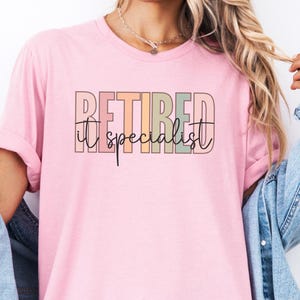 May include: A light pink t-shirt with the words "RETIRED IT specialist" printed across the front in a colorful, retro font. The text is in shades of pink, green, orange, and black.