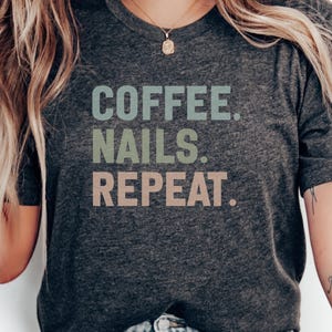 May include: A dark grey t-shirt featuring the text "COFFEE. NAILS. REPEAT." in pastel green, blue, and pink. The top has a crew neck and short sleeves.