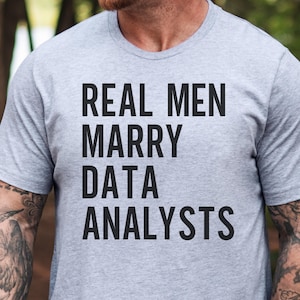 May include: A heather grey short-sleeved t-shirt featuring the black text "REAL MEN MARRY DATA ANALYSTS" printed in a bold, sans-serif font across the chest. The garment has a classic crew neckline and a standard fit, showcasing a simple design.