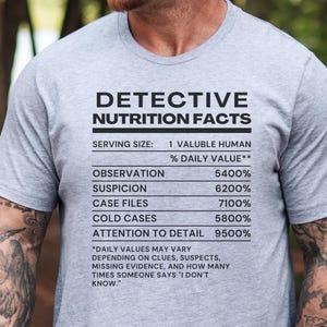 May include: A heather grey short-sleeve t-shirt featuring black text that reads: DETECTIVE NUTRITION FACTS, SERVING SIZE: 1 VALUABLE HUMAN, % DAILY VALUE, OBSERVATION 5400%, SUSPICION 6200%, CASE FILES 7100%, COLD CASES 5800%, ATTENTION TO DETAIL 9500%.