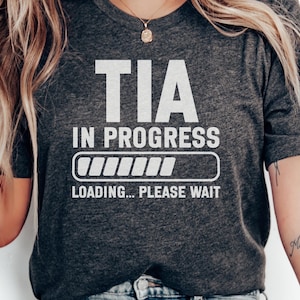 May include: A dark heather grey t-shirt featuring white text that reads TIA IN PROGRESS with a loading bar graphic below it and the text LOADING... PLEASE WAIT underneath the bar. The shirt has short sleeves and a crew neckline.