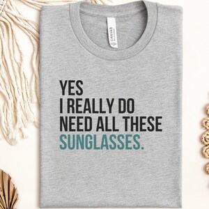 May include: A heather grey short-sleeved t-shirt with a crew neckline. The front features black text that reads YES I REALLY DO NEED ALL THESE and the word SUNGLASSES in teal. A white brand label is visible inside the collar of the garment.