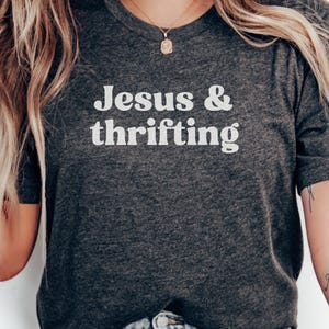 May include: Dark gray t-shirt with the words "Jesus & thrifting" in white block letters. The shirt is a crew neck style. A gold necklace with a pendant is visible.