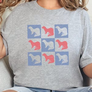 Puede incluir: Camiseta gris claro con un patrón repetido de hurones rojos y blancos dentro de cuadrados azules. La camiseta tiene mangas cortas y cuello redondo.
