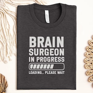 May include: Charcoal grey t-shirt featuring the white text "BRAIN SURGEON IN PROGRESS", a loading bar graphic, and the words "LOADING... PLEASE WAIT." The shirt is folded and rests on a neutral surface.